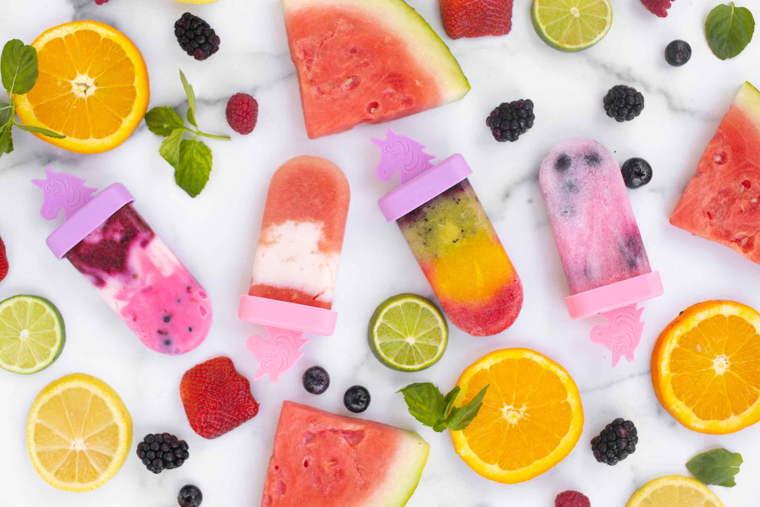 Lifestyle image of rainbow and unicorns ice pops surrounded by various fruits.