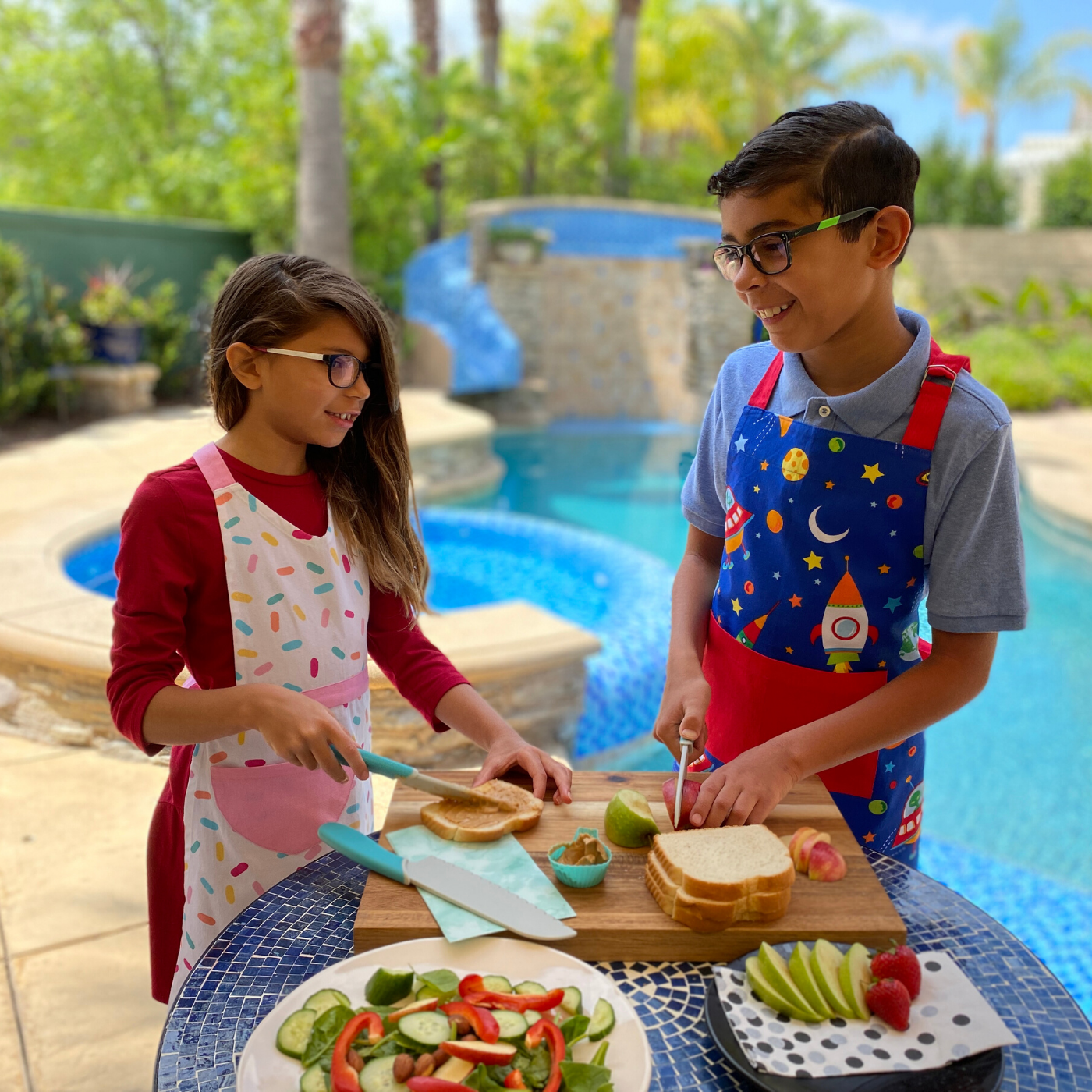 Lifestyle image of Chopping Block Kid Safe Nylon Serrated 3 Pack Knife Set being used to make a peanut butter and jelly sandwich as well as slicing apples