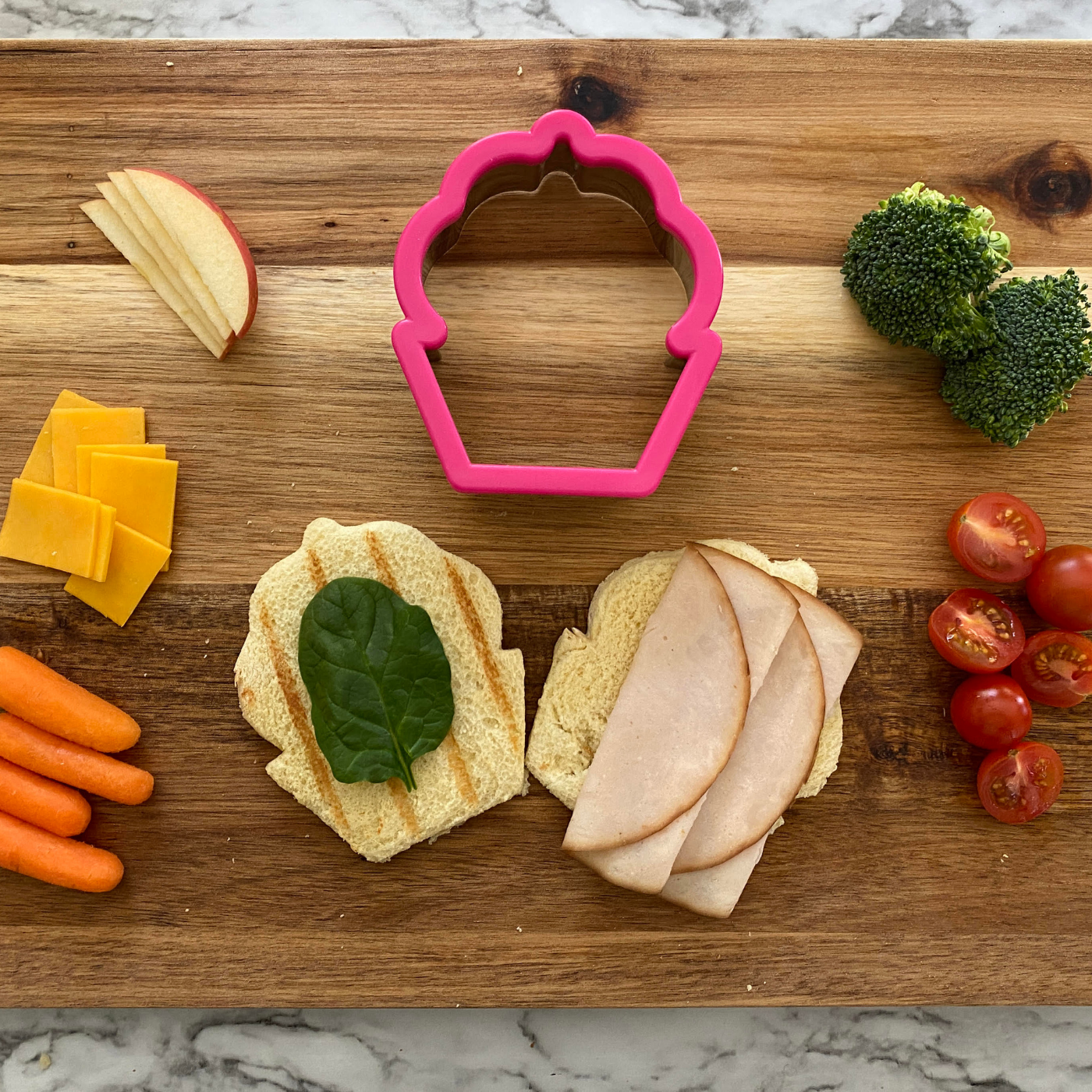 Lifestyle photo of stainless steel cupcake shaped cookie cutter along with meat, cheese, and vegetables to assemble a cupcake shaped sandwich.