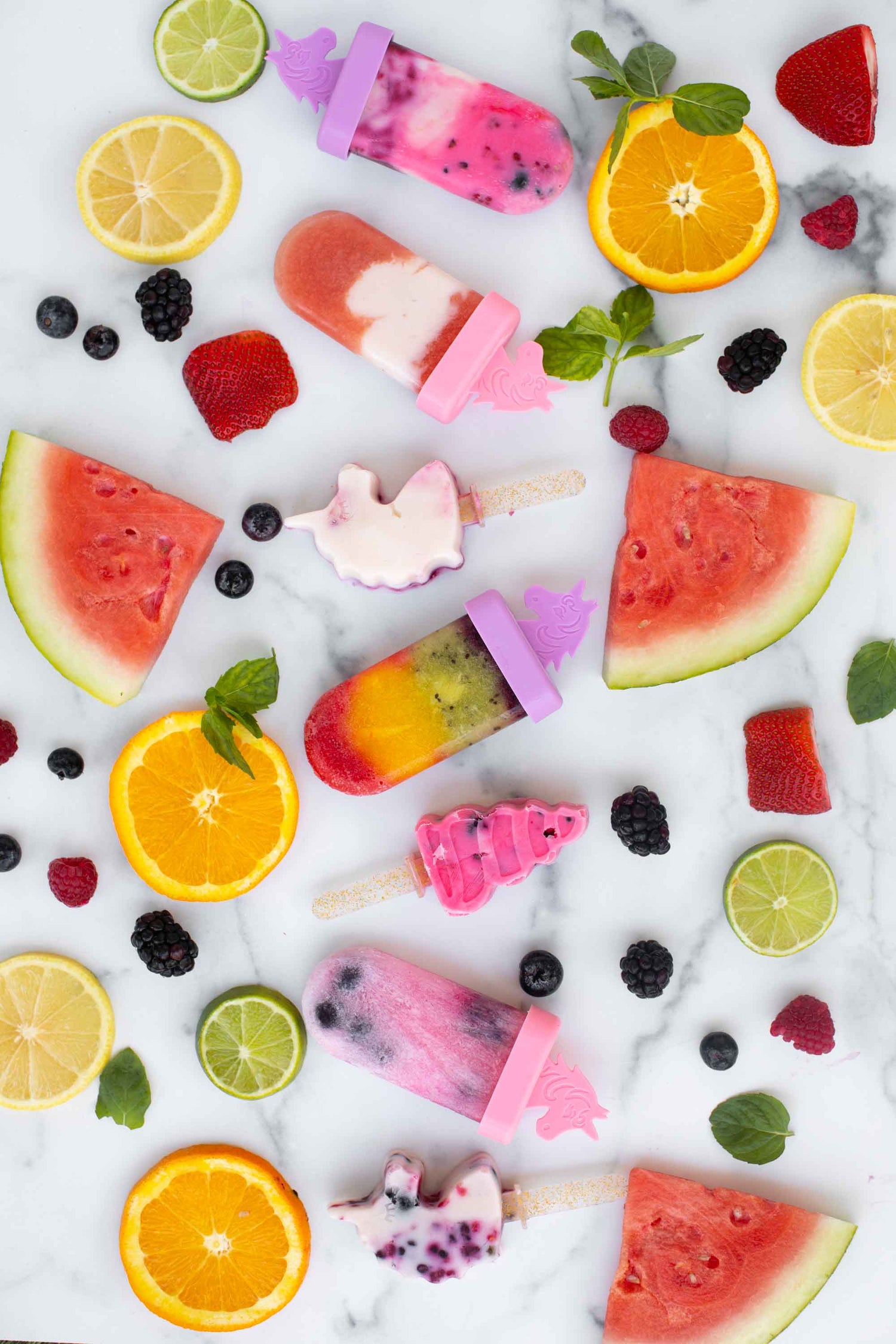 Lifestyle image of rainbow and unicorns ice pops surrounded by various fruits.  Edit alt text