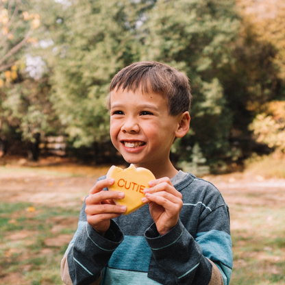 Lifestyle image of young boy eating yellow heart shaped cookie that says &quot;cutie&quot;