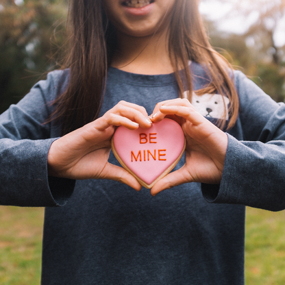 lifestyle photo of girl holding a pink heart shaped cookie that says &quot;be mine&quot; 