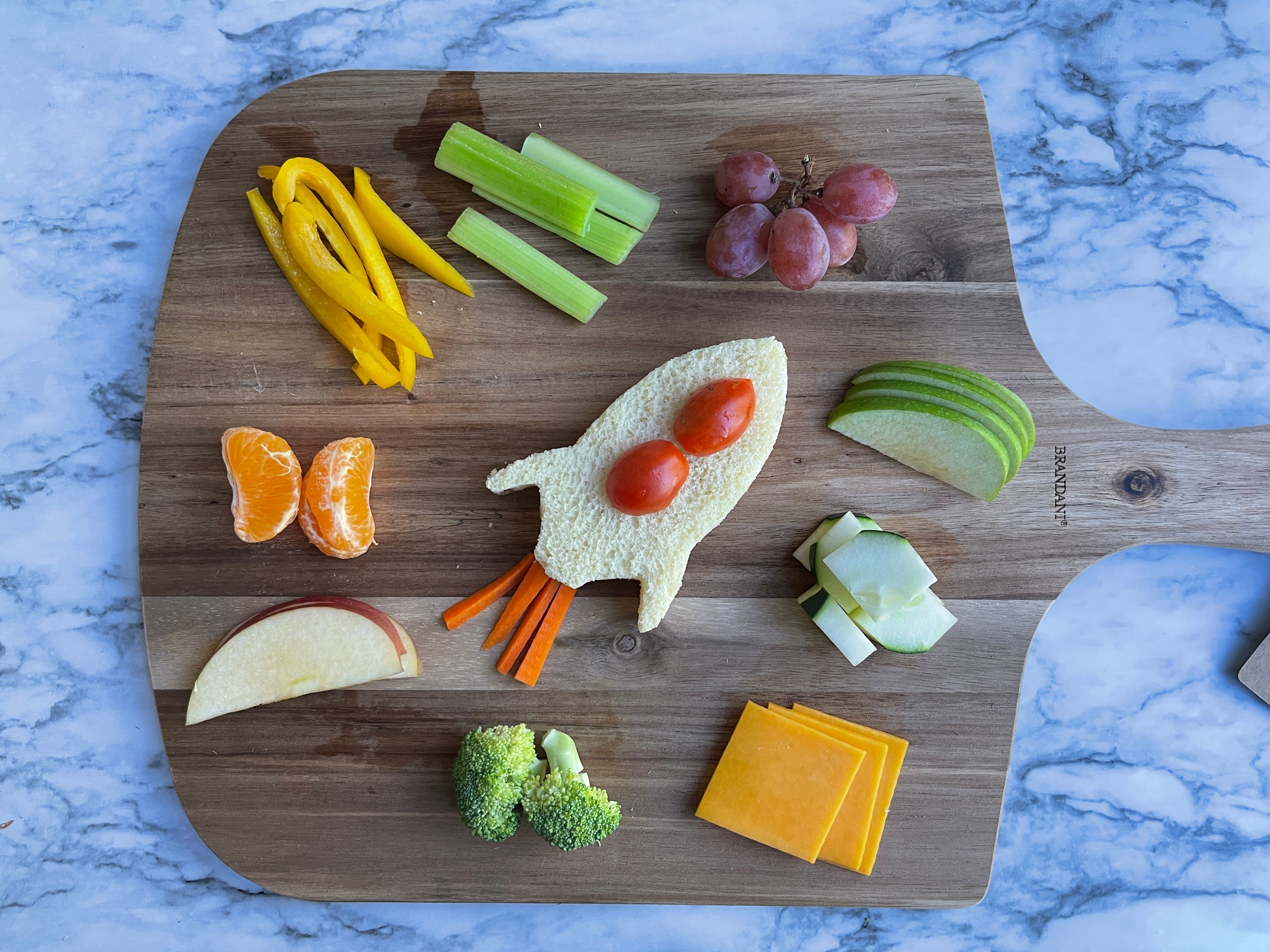 Lifestyle image of a rocket ship shaped sandwich on a cutting board surrounded by fruits and vegetables 