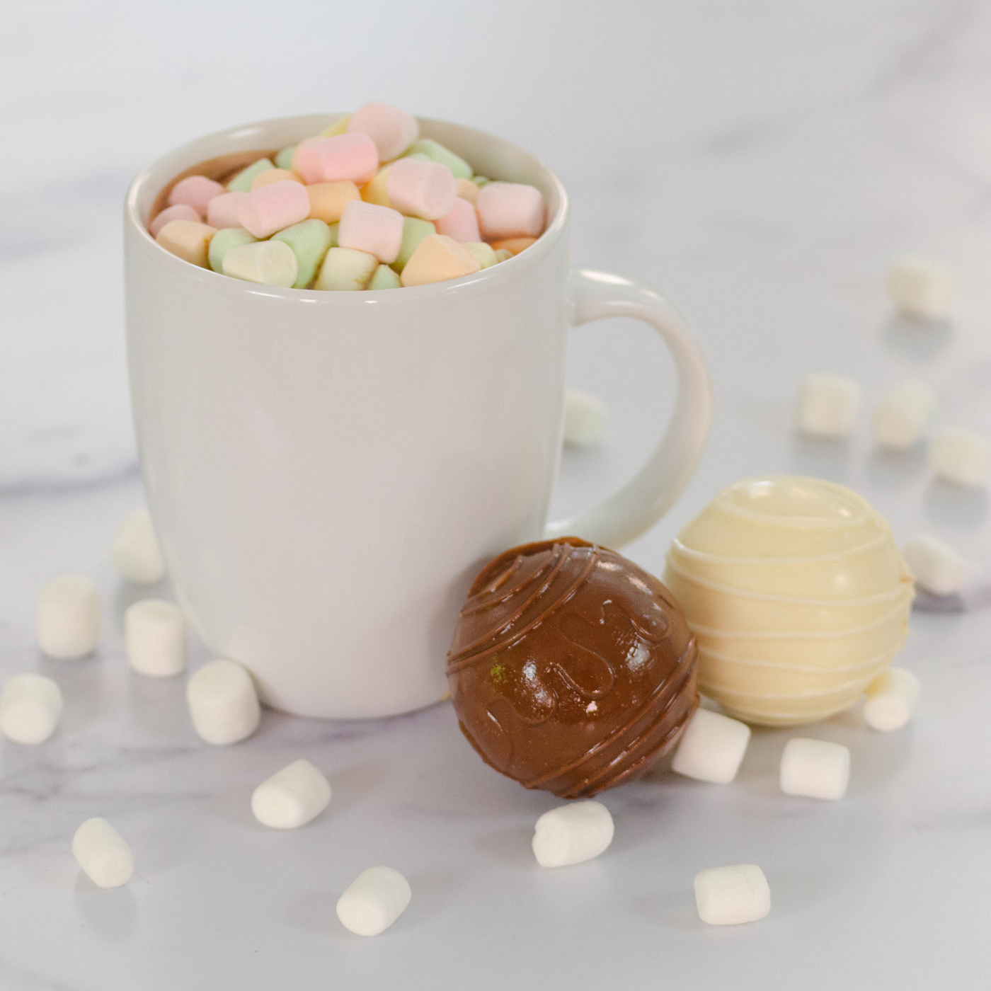 Lifestyle image of two hot cocoa bombs alongside a mug of hot chocolate with marshmellows