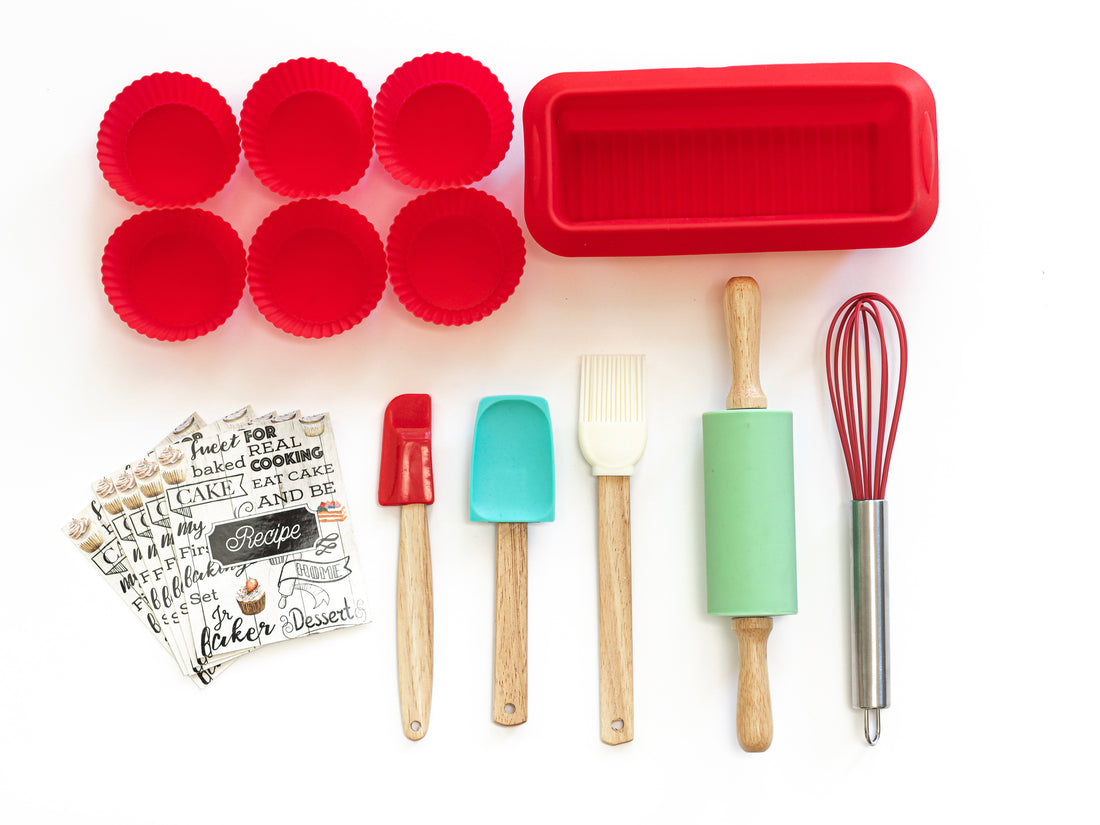 Out of box image of Junior baker baking set including:1 spatula, 1 pastry brush, 1 mixing spoon, 1 silicone loaf pan, 6 silicone baking cups, 1 rolling pin, 1 whisk, and 5 recipe cards.