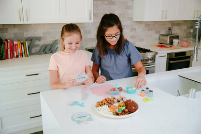 Lifestyle image of two girls decorating the Rainbows &amp; Unicorns CHARcuteRIE Set with various snacks.