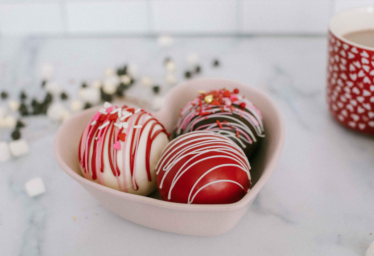 Lifestyle image of three valentines themed hot cocoa bombs in a heart shaped bowl