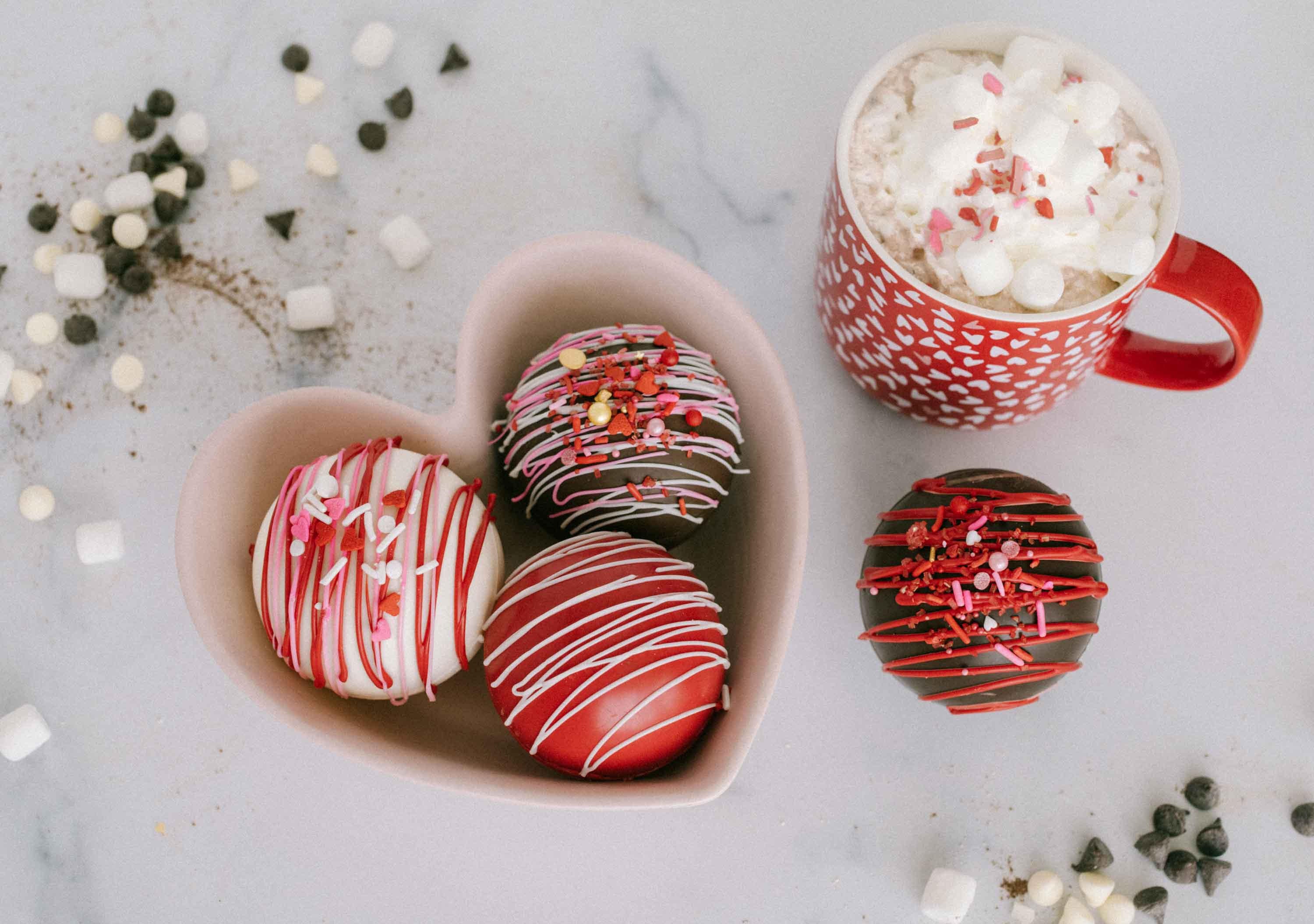 Lifestyle image of four valentines themed hot cocoa bombs in a heart shaped bowl